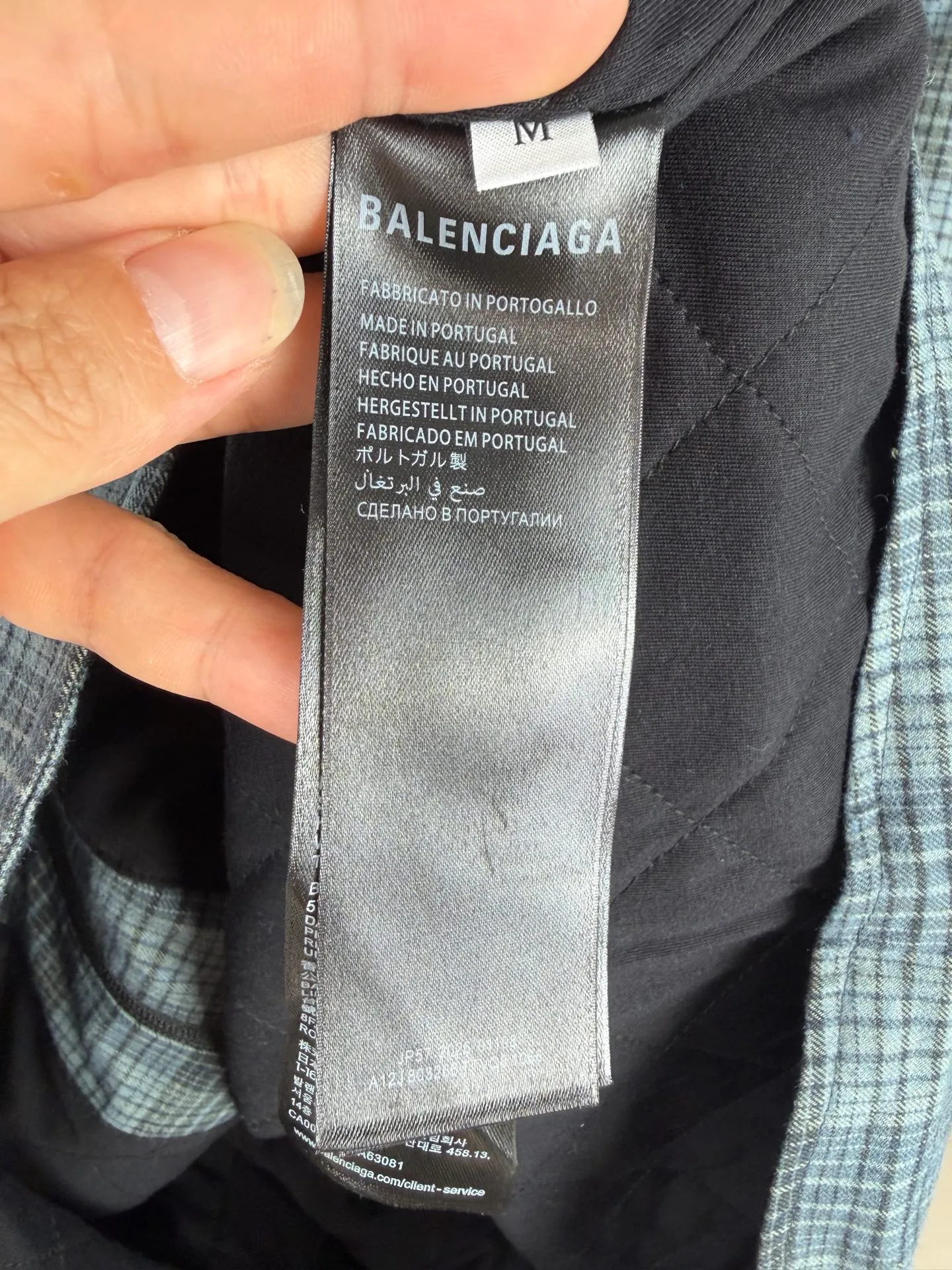 BALENCIAGA/balenciaga blue and gray plaid zipper pocket logo quilted jacket combines a three-layer structure of plaid woven fabric, filling cotton, and lining, and integrates plaid alignment technology and quilted garment production standards. There are multiple process difficulties in the whole process. The surface layer of the body is made of blue-gray Retro plaid fabric, and the cutting machine production strictly follows the global grid standards. The arrangement direction of the cutting pieces is fixed and cannot be changed at will. The horizontal and vertical lines of shoulder seams, side seams, and zipper placket joints must be consistent and coherent. The left chest patch pocket is cut independently, and the bag check pattern must be aligned with the front reference pattern. Slight deviations will destroy the visual integrity and directly increase fabric loss and cutting time. The most difficult part of the style is the three-layer composite sewing. The outer plaid fabric, filling cotton, and lining fabric have different physical properties, and the preshrinking processing parameters need to be adjusted separately. Pressure and temperature control are crucial during lamination operations. Once the parameters are out of balance, it can easily cause fabric stretch and distortion, plaid dislocation, and wrinkles and bulges in garments. At the same time, it is necessary to reasonably plan the quilting line to avoid the visual beauty of the grid pattern from being cut by the lines on the basis of firmly fixing the cotton layer and preventing cotton from running away. The placket adopts a centered zipper structure. It is difficult to sew the zipper tape and the quilted panels. It is required that the width of the left and right plackets be equal, the zipper routing is straight and smooth, and there is no lag in opening and closing. When the shirt lapel is filled with cotton, the thickness of the cotton needs to be controlled to prevent the collar from becoming bloated and unnaturally folded. The metal nameplate of the chest pocket is sewn at fixed points to avoid pulling the fabric during the operation and ensure that the plaid pattern is smooth and not deformed. The quilted material increases the overall thickness of the fabric. When sewing, you need to continuously adjust the presser foot pressure and stitch length to adapt to changes in sandwich thickness. The low-temperature and gentle ironing process is used for post-finishing to prevent the plaid from fading and the cotton layer from clumping due to high temperatures. From the precise alignment of cutting pieces, three-layer fabric pretreatment, composite quilting to garment finalization inspection, the processes are interlocking and the error tolerance rate is extremely low, testing the accuracy of plate making, equipment control and turning technology. Size:XS S M L